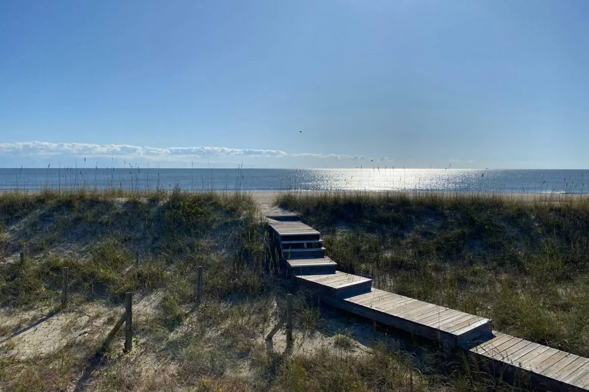 A wide wooden pathway leads through sand dunes with sea grasses toward an open ocean view. The morning light creates a serene, inviting mood, and the path suggests easy access for families headed to the beach for play or exploration.