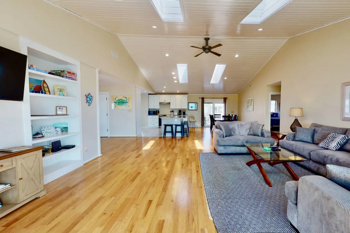 A spacious, light-filled living area with comfortable seating, an open kitchen visible in the background, and natural light pouring in from skylights. The space feels airy and inviting, perfect for gatherings and family activities.