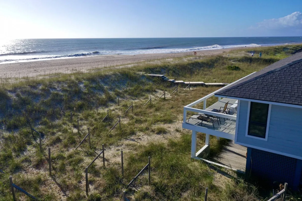 A beachfront deck furnished with inviting lounge chairs and a table, overlooking rolling ocean waves. The sun is either rising or setting, bathing the deck in warm light. The deck appears spacious, with ample room for relaxing and dining while enjoying panoramic ocean views.