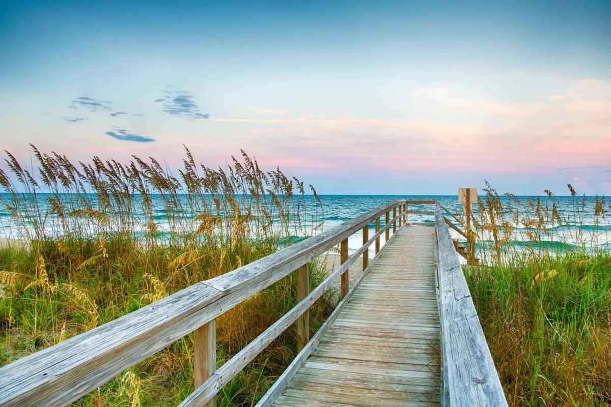 A serene exterior scene featuring a wooden boardwalk flanked by dune grass, leading out to the ocean. Activities like walking and beachcombing are suggested by the open pathway, and the tranquil mood makes it ideal for sunset viewing and peaceful strolls.