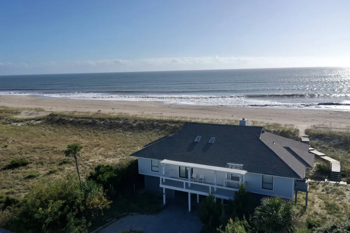 A view of the beachfront property framed by lush vegetation, with gentle waves rolling in. The expansive shore is perfect for families and pets to enjoy activities like beachcombing, sunbathing, and evening walks together.
