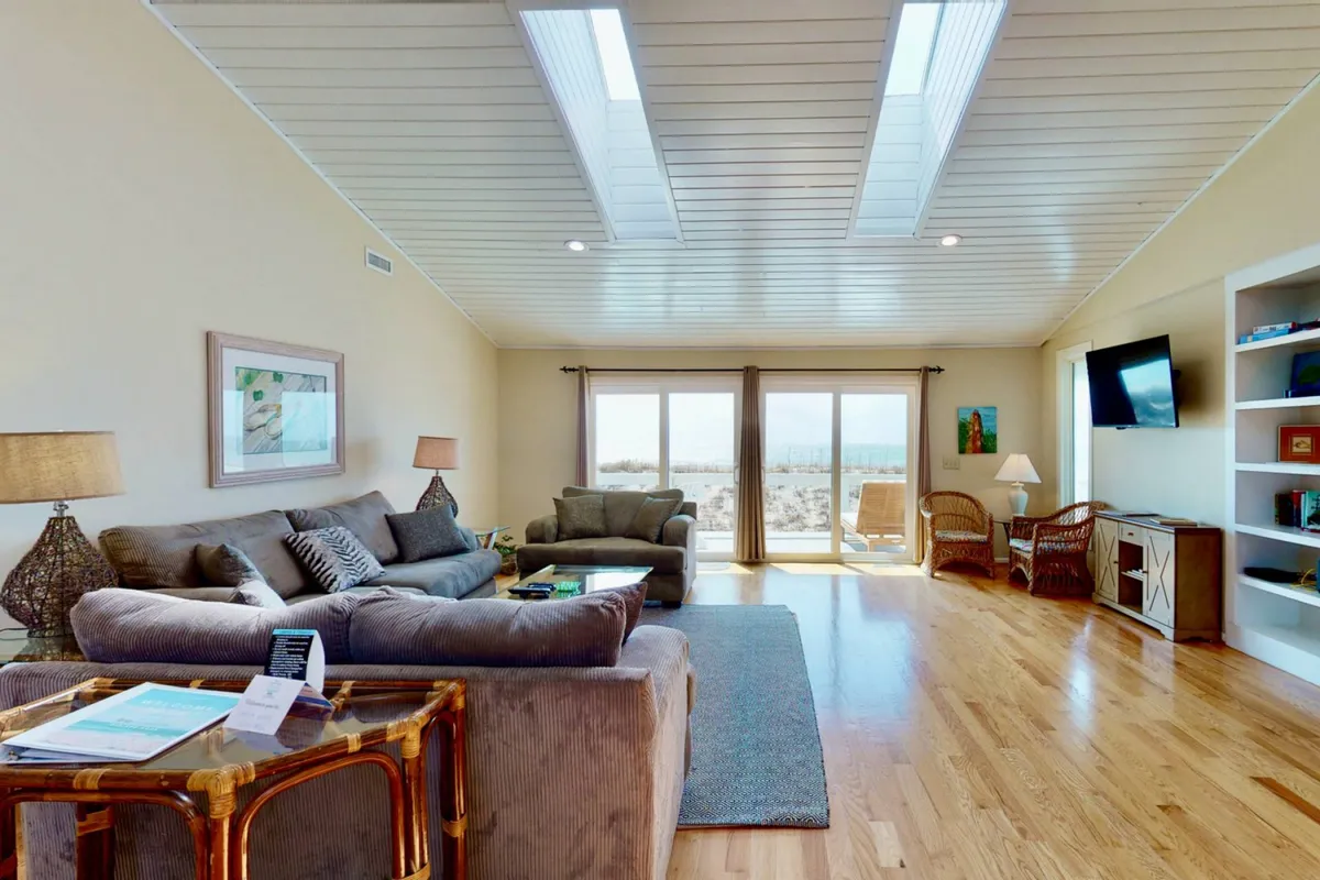 A welcoming living room with plush sofas, vaulted ceilings, large panoramic windows, skylights, an entertainment unit, and coastal decor. The room is designed for comfort, with natural light enhancing the breezy vibe.