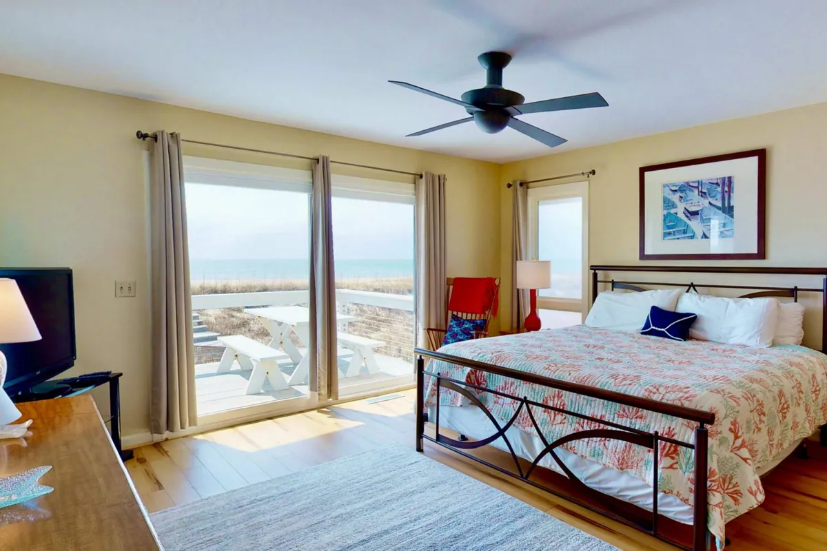 A beautifully appointed bedroom featuring a plush bed with patterned bedding, a colorful armchair, and large windows that offer sweeping views of the beach. The tranquil atmosphere is enhanced by the natural light pouring in, giving the room a cozy and inviting feel.