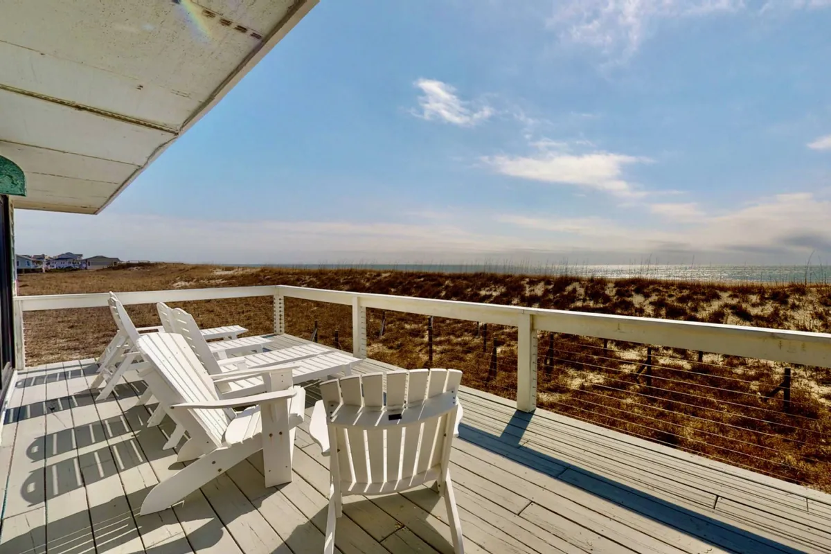 A wooden deck furnished with Adirondack chairs arranged to face an unobstructed, sweeping view of the ocean and dunes. The setting invites relaxation, sunset viewing, and social gatherings, capturing the tranquil ambiance of outdoor living.
