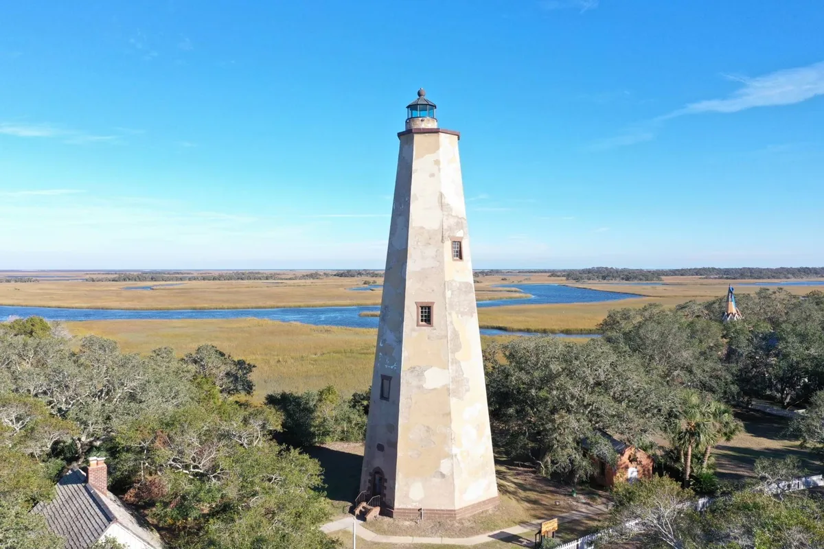 This image features the iconic Bald Head Island lighthouse surrounded by marshland and a peaceful river, with trees framing the scene. The lighthouse stands tall at the center, evoking a sense of adventure, exploration, and island history.