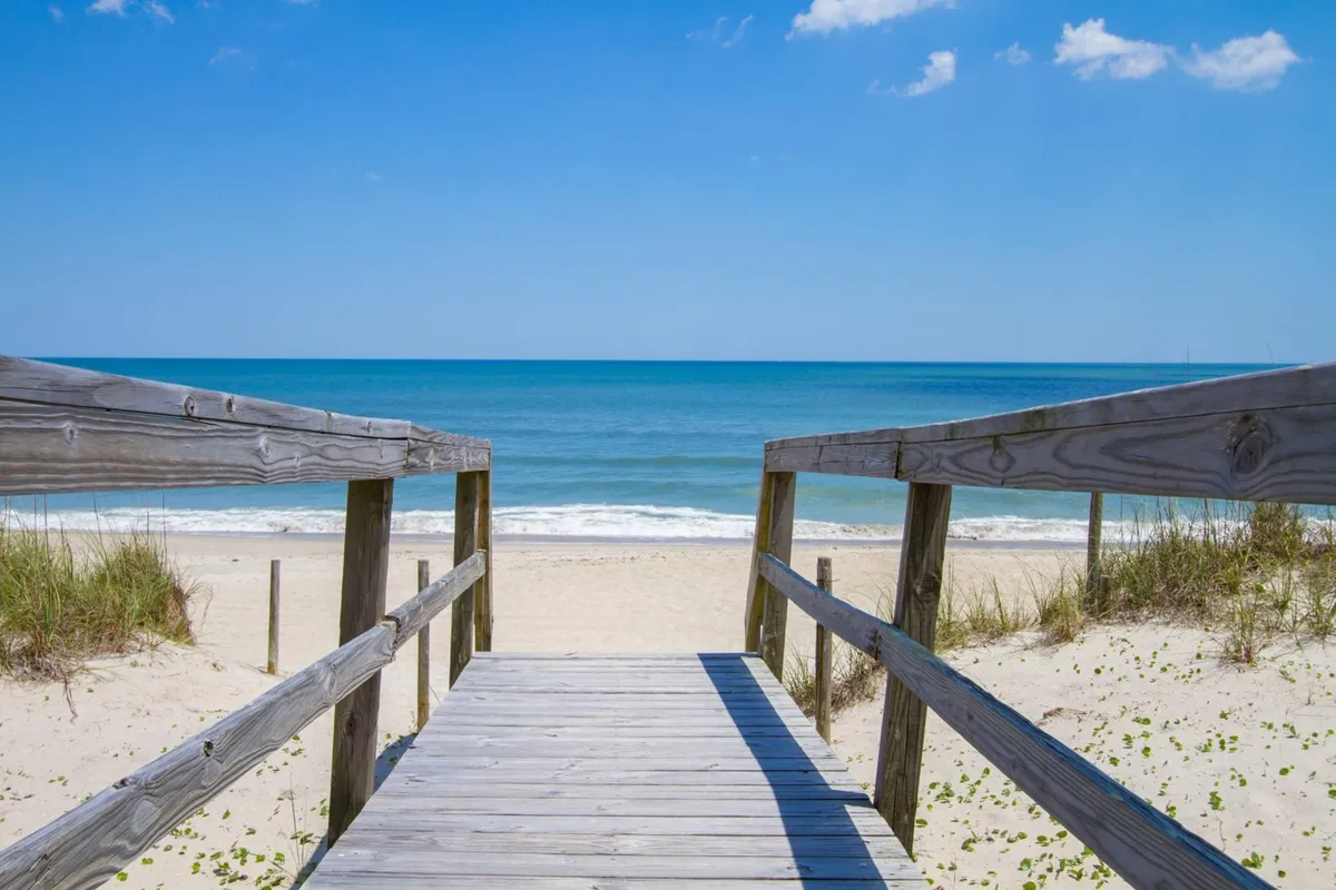 A wooden walkway leads over dune grass to a sandy beach with the ocean visible in the background. The scene conveys a sense of relaxation and openness, ideal for leisurely strolls and enjoying the outdoors. The warm lighting suggests a late afternoon or sunset walk.