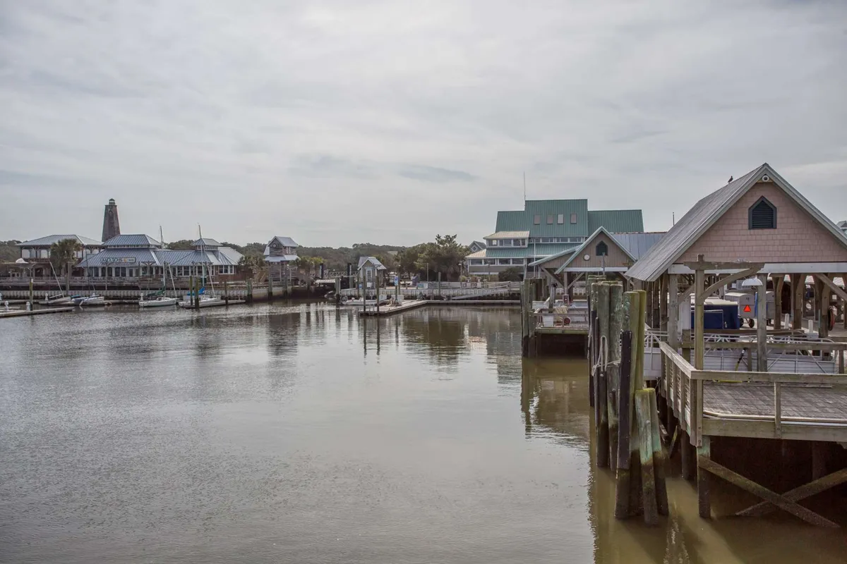 A vibrant view of waterfront docks filled with boats, quaint buildings, and the lighthouse visible in the distance. The marina is bustling with possibilities for boating, fishing, and exploring the island by water.