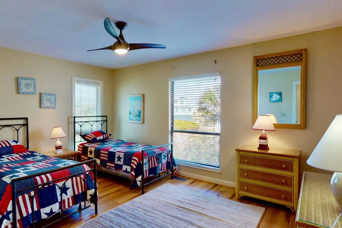 A comfortable twin bedroom with two beds dressed in patriotic quilts, a window with blinds, and warm lamplight, creating a welcoming and child-friendly space. Decorative touches and a mirror add to the homely feel, perfect for younger guests.