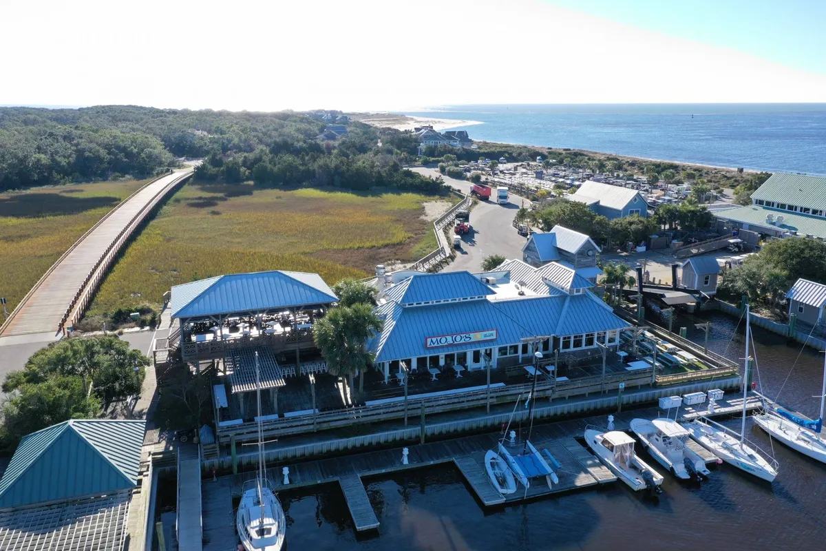 This relaxed, vibrant exterior image centers on a marina-side restaurant, with boats docked nearby and lush landscaping. The atmosphere is lively yet peaceful, with clear opportunities for waterfront dining and sunset views from the deck.